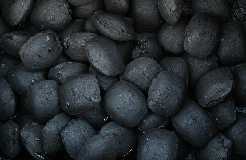 A pile of dark, irregularly shaped briquettes, likely industrial or mineral in nature, with several pairs of shoes visible around, indicating a work or inspection scene.
